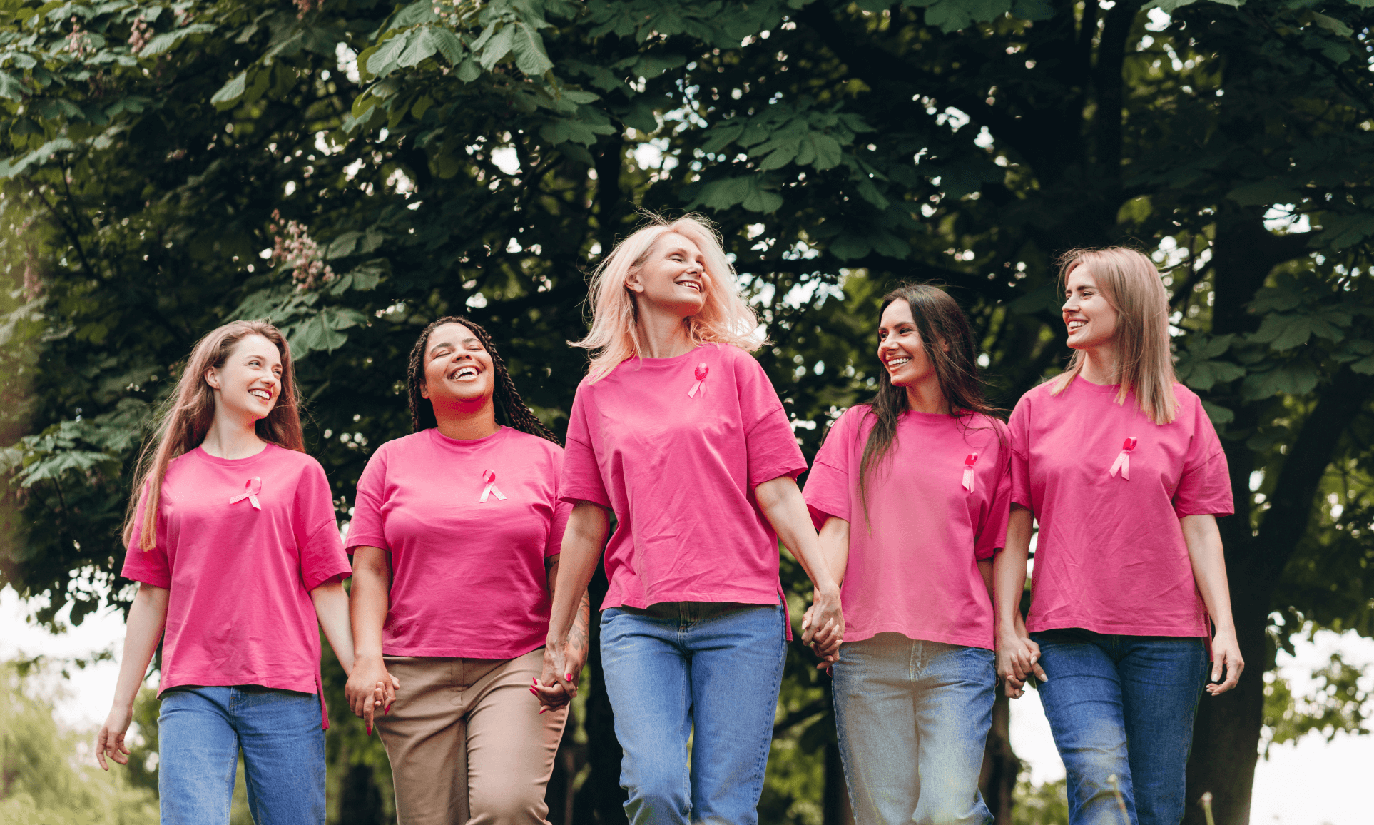 Donne unite per la prevenzione del tumore al seno – Campagna Ottobre Rosa. Cinque donne sorridenti camminano mano nella mano in un parco, indossando magliette rosa con il nastro simbolo della prevenzione del tumore al seno, durante un evento di sensibilizzazione per l’Ottobre Rosa.