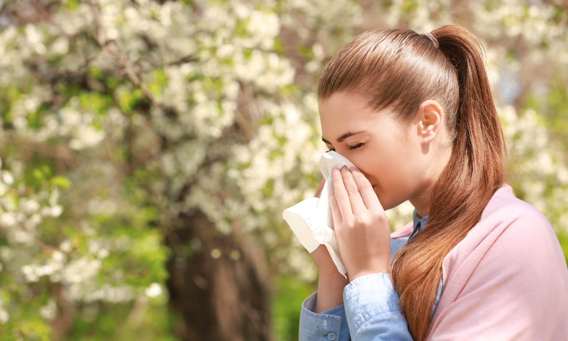 Allergie stagionali: come proteggersi fuori e dentro casa Donna all’aperto tra alberi in fiore che si soffia il naso con un fazzoletto, sintomo delle allergie stagionali primaverili.